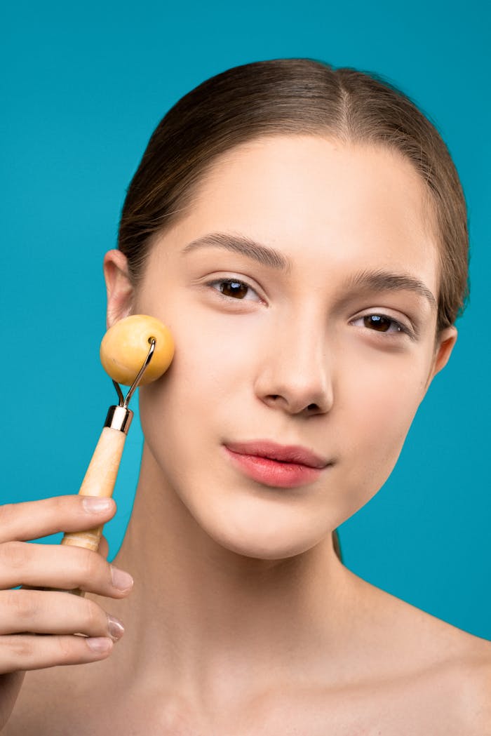 A woman smiling while using a wooden facial roller for skin care on a blue background.