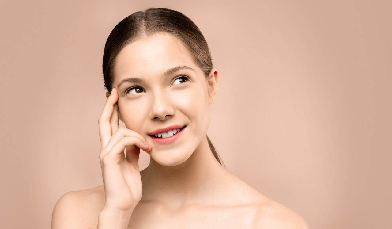 A young woman with flawless skin gazes thoughtfully upwards against a neutral background.